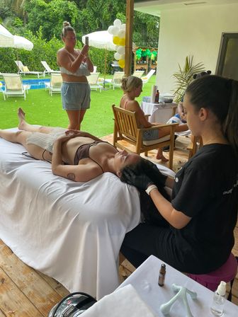 Poolside spa scene in a tropical backyard: a person reclines on a white-covered massage table while a therapist performs a head massage, with guests on wooden chairs, balloons and sun loungers beside a blue pool.