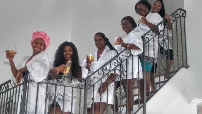 Six women in white satin robes pose on a decorative wrought-iron indoor staircase, smiling and holding orange cocktails in a cheerful getting-ready celebration.