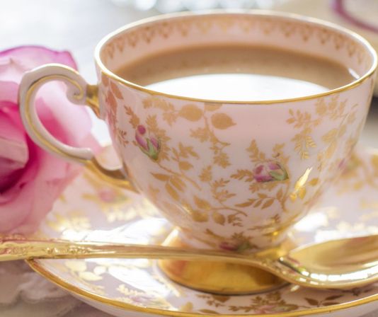Vintage porcelain teacup with pink rose motifs and gold filigree, filled with tea on a matching saucer with a gold teaspoon