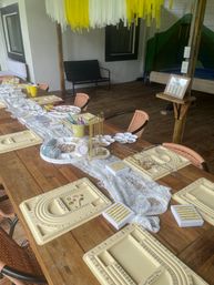 Outdoor covered patio set up for a DIY jewelry-making party: long wooden table with bead design boards, trays of chains and beads, tools, lace runner, wicker chairs and yellow hanging decorations.
