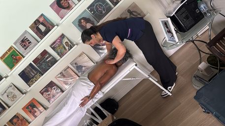 Massage therapist giving a shoulder massage to a client lying face-down on a portable table in a cozy home studio with a wall of vinyl records and hardwood floors.