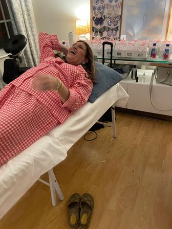 Laughing person in red gingham pajamas reclining on a massage table in a cozy home-spa room with butterfly wall art, pink gift boxes on a shelf and slippers on a wood floor.