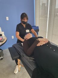 Esthetician in black scrubs and mask giving a relaxing facial with cooling eye pads to a client reclined on a treatment table in a modern blue-walled spa room.