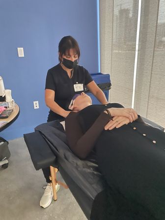 Esthetician in black scrubs and mask giving a relaxing facial with cooling eye pads to a client reclined on a treatment table in a modern blue-walled spa room.