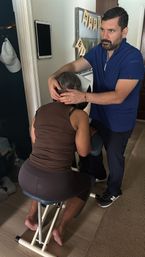 In-home chair massage: therapist in blue scrubs adjusting a client’s neck on a portable seated massage chair in a residential room, calm wellness session.