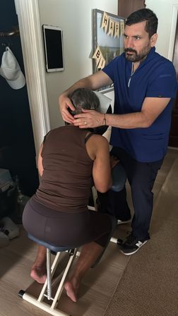 In-home chair massage: therapist in blue scrubs adjusting a client’s neck on a portable seated massage chair in a residential room, calm wellness session.