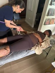 Client reclining on a massage table wearing an eye mask as a therapist performs a hand and leg massage in a cozy indoor spa studio with vinyl artwork on the wall.