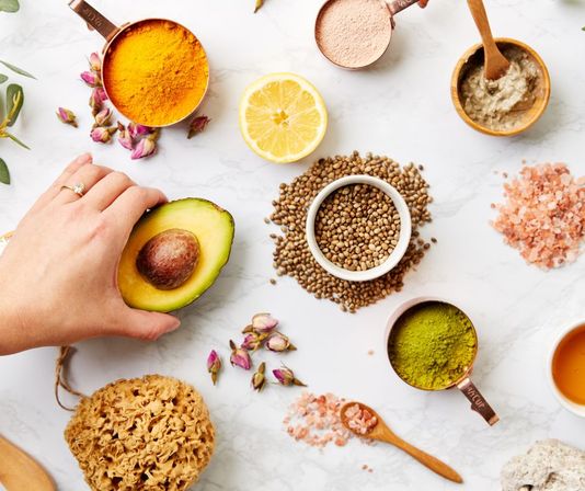 Bright flatlay of natural skincare ingredients on white marble: hand holding halved avocado, turmeric and matcha powders, hemp seeds, pink Himalayan salt, lemon half, dried rosebuds, clay mask and sea sponge