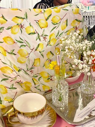 Cheerful lemon-print tablecloth with baby's-breath flowers, crystal perfume bottle and gold-rim teacup arranged on a mirrored tray — bright summer brunch tablescape.