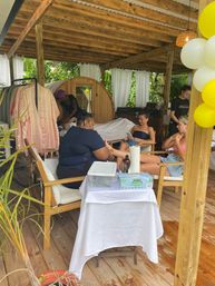 Tropical outdoor cabana spa with wooden pergola and barrel sauna, spa staff giving pedicures and massages to relaxed clients on lounge chairs under white curtains and yellow balloons.