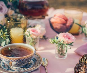 Ornate blue-patterned teacup of tea on a saucer with a gold spoon, surrounded by small pink rose bud vases, amber glasses and pastries on a pastel pink tablecloth in a sunny garden tea-party setup.
