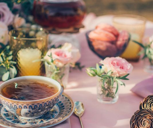 Ornate blue-patterned teacup of tea on a saucer with a gold spoon, surrounded by small pink rose bud vases, amber glasses and pastries on a pastel pink tablecloth in a sunny garden tea-party setup.