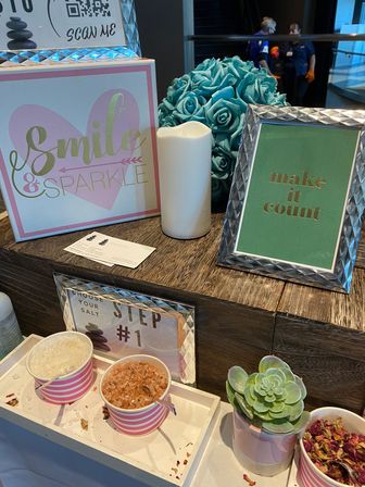 Cheerful spa counter display with pink-striped bowls of bath salts, potted succulent, teal rose bouquet, LED candle and framed signs reading "Smile & Sparkle" and "make it count".