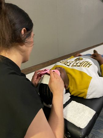 Practitioner places a pink eye mask on a client lying on a treatment table in a spa/skincare setting; client wears a yellow sweatshirt and towels cushion the head.