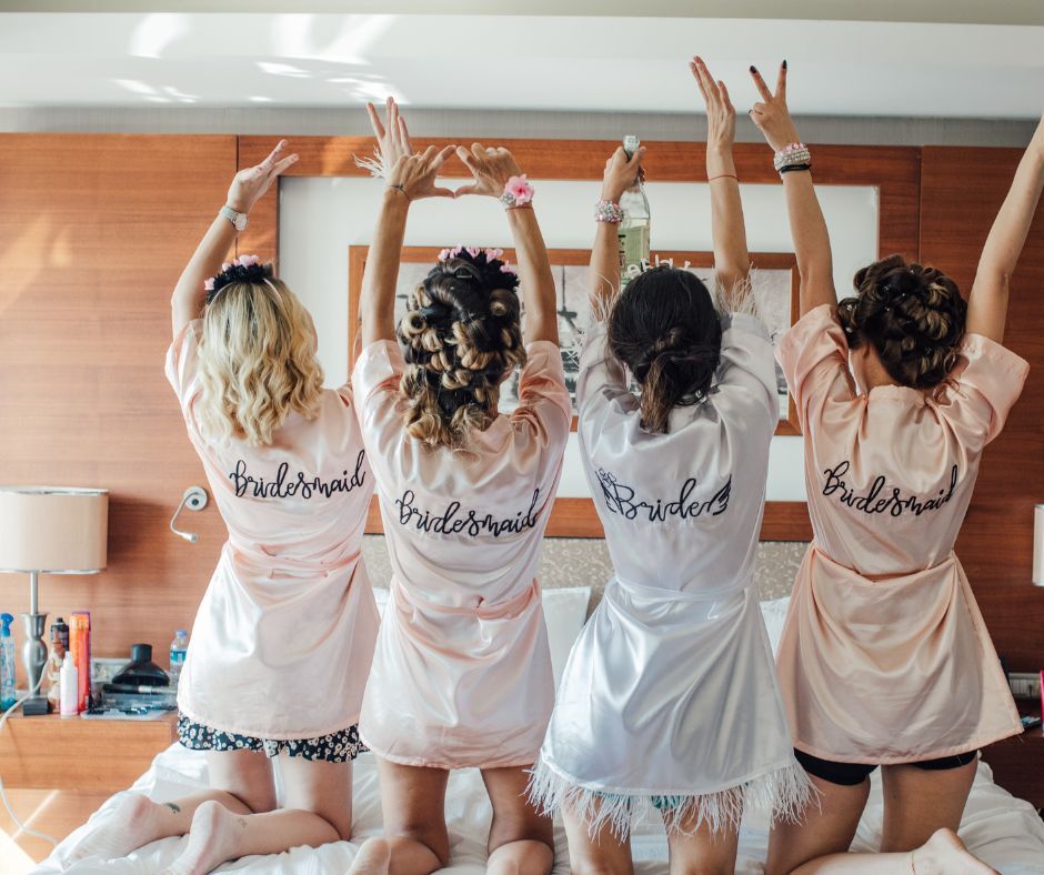 Four women kneeling on a hotel bed in satin robes — three blush ‘Bridesmaid’ robes and one white ‘Bride’ robe with feather trim — arms raised in a celebratory wedding-morning getting-ready scene.