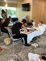 Group pedicure and pampering in a waterfront boat lounge, women reclining on spa chairs receiving foot treatments with windows overlooking a canal