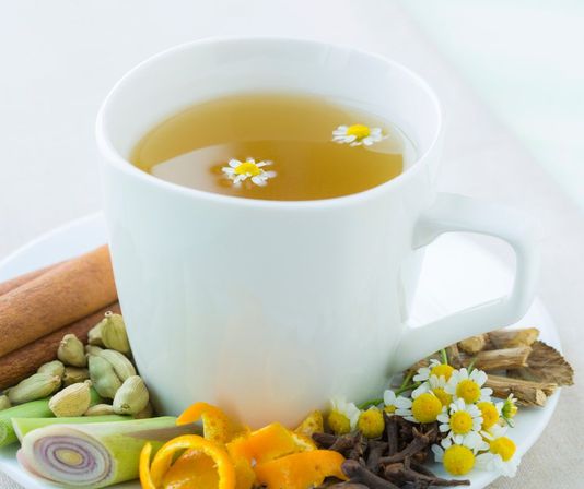 Soothing white mug of chamomile herbal tea with two floating chamomile flowers, on a saucer surrounded by cinnamon sticks, cardamom pods, lemongrass, orange peel curls, cloves, and fresh chamomile blossoms.