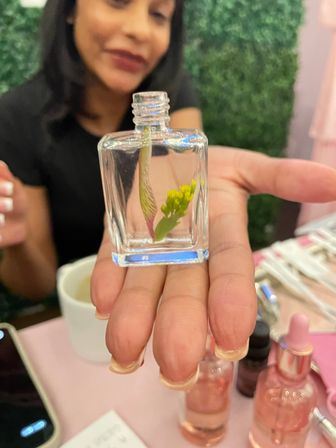 Close-up of a hand holding a tiny square glass bottle with a small yellow flower stem inside, pink beauty bottles and a blurred person in the background.