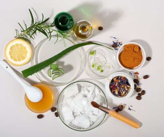 DIY natural skincare flat lay on white: aloe vera leaf and gel, lemon half, rosemary sprig, glass bowls with coconut flakes, honey, oil, turmeric powder, dried flower petals, scattered coffee beans and a makeup brush — organic beauty ingredients.