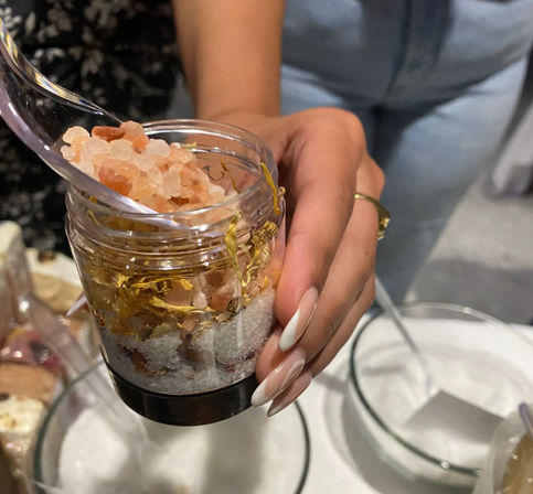 Close-up of a hand with white almond nails holding a jar of artisan bath salts layered with pink Himalayan crystals, white sea salt and yellow dried flower petals, clear scoop and glass bowls in the background.