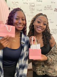 Two smiling women holding small pink gift bags in front of a wall of inspirational quotes — cheerful friends enjoying a self-care gift moment