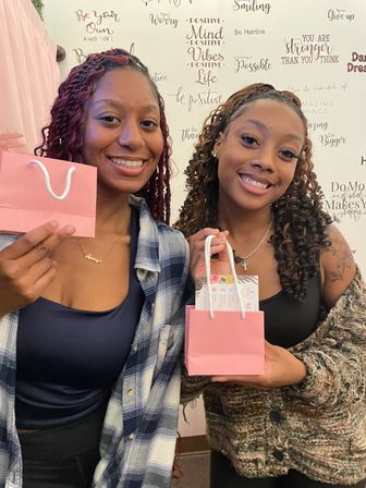 Two smiling women holding small pink gift bags in front of a wall of inspirational quotes — cheerful friends enjoying a self-care gift moment
