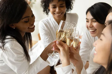 Four women in white robes smiling and clinking champagne flutes in a bright indoor celebration, a joyful toast among friends.