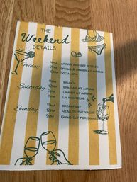 Yellow-and-white striped weekend itinerary titled 'The Weekend Details' on a wooden table — lists Friday, Saturday and Sunday with times and activities (arrive, drinks and dinner, brunch, spa day, nightclub, yacht outing) and playful cocktail, wine and bikini illustrations.