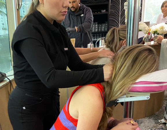 Seated chair massage with a therapist in black working on a woman in a red and purple striped top at an indoor salon or spa counter