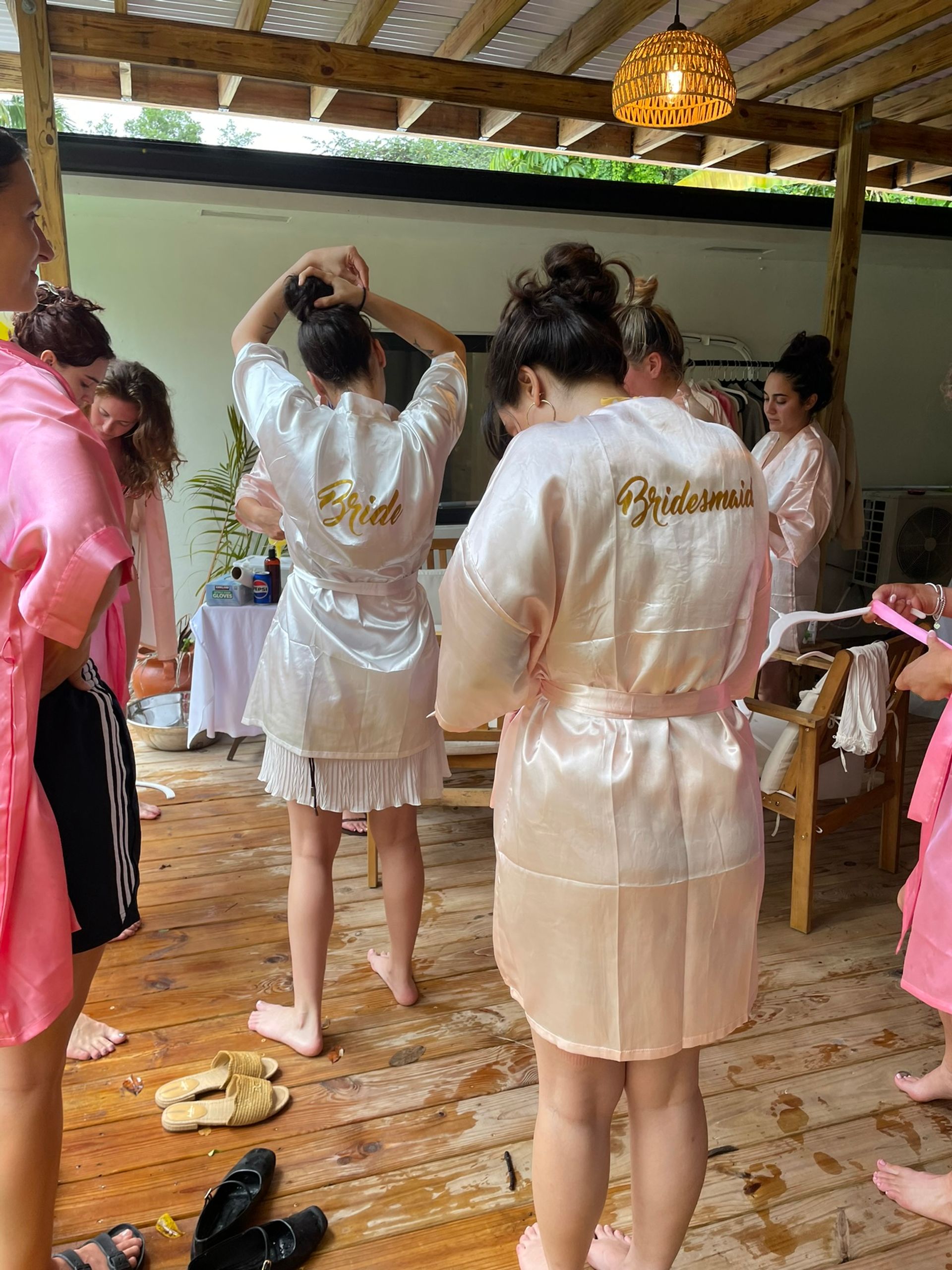 Bridal party getting ready on a wooden deck under a covered patio — women in matching satin robes with gold “Bride” and “Bridesmaid” lettering fixing hair and prepping.