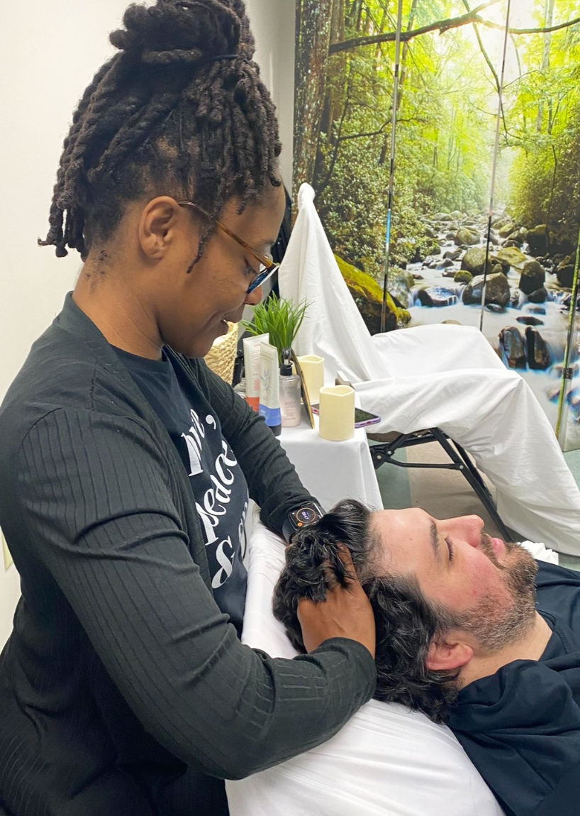 Spa therapist giving a relaxing scalp massage to a man reclining on a treatment table in a wellness room with a forest stream mural, candles and skincare products nearby.