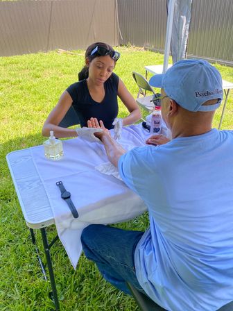 Therapist giving a relaxing outdoor hand massage to a seated man at a white-covered folding table on a sunny backyard lawn, with a lotion pump, smartwatch and drink bottle visible under a canopy.