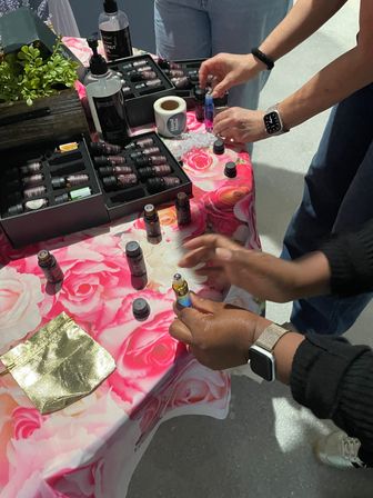Hands sampling roll-on essential oils and small aromatherapy bottles at an indoor vendor table with display cases on a pink rose-patterned tablecloth — wellness product sampling.