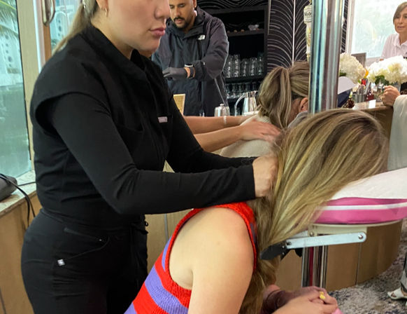 Therapist giving a seated chair neck and shoulder massage to a woman on a portable face-cradle chair at an indoor café-style wellness event, with a bar counter and other attendees in the background.