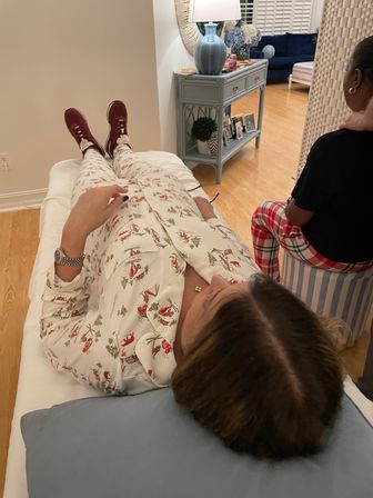 Person in holiday-print pajamas reclining on a couch with maroon sneakers and a blue pillow, cozy living room scene with a blue side table, lamp and framed photos, and a companion seated nearby in red plaid pants.