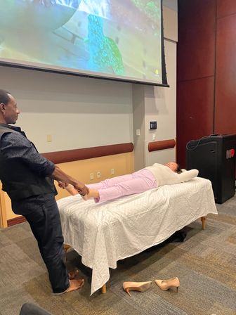 Wellness workshop in a seminar room: participant reclines on a white-sheeted portable massage table while an instructor demonstrates foot reflexology in front of a large projection screen; beige heels on the carpet.