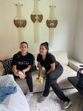 Two women sitting on a beige sofa in a cozy living room, wearing casual black outfits and sneakers and snacking from chip bags; geometric throw pillow and three macramé wall hangings decorate the neutral wall.