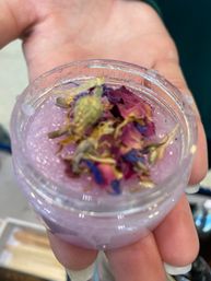 Hand holding clear jar of pink floral sugar scrub topped with dried rose and blue cornflower petals — handmade spa exfoliant