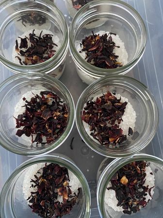 Top-down view of seven clear glass jars on a plastic tray, each filled with coarse white bath salts and topped with dark red dried hibiscus petals — DIY floral bath soak for spa or gift.