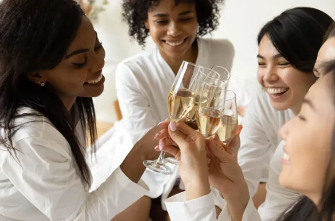 Close-up of friends in white robes clinking champagne flutes and smiling during an indoor celebratory toast.