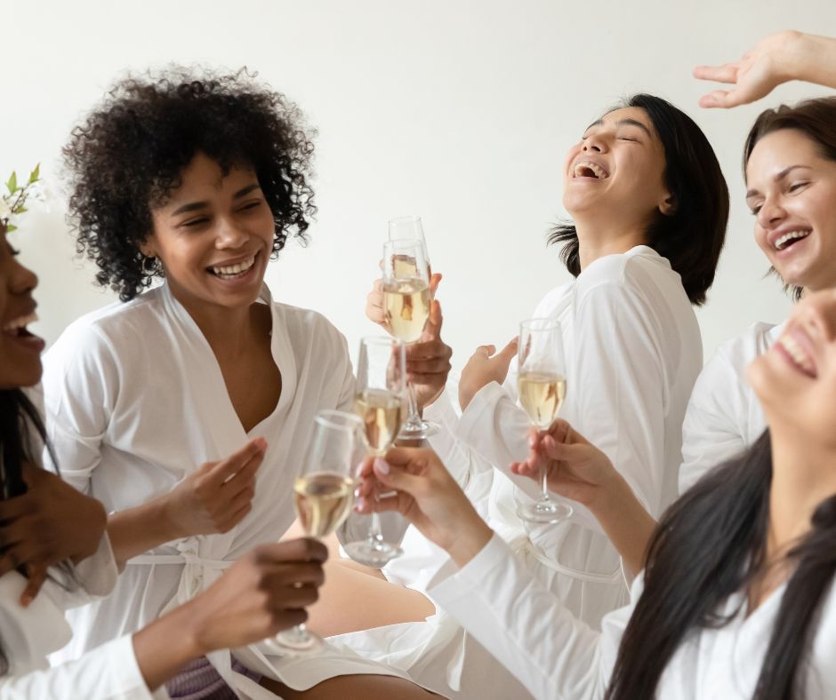 Group of women in white robes laughing and toasting champagne in a bright, sunlit indoor spa-like setting — fun girls’ night celebration.