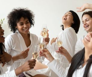 Group of women in white robes laughing and toasting champagne in a bright, sunlit indoor spa-like setting — fun girls’ night celebration.