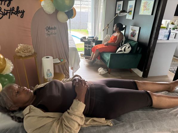 Home spa birthday scene in a living room: balloons and floral decor by a treatment table with a woman lying relaxed, while another guest in an orange dress soaks her feet on a green sofa near sliding doors overlooking a backyard pool.