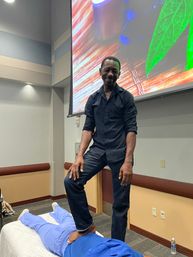 Practitioner demonstrating a standing foot-on-back massage on a person lying face down during a wellness workshop in a conference room with a large colorful projection screen.