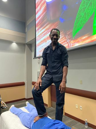 Practitioner demonstrating a standing foot-on-back massage on a person lying face down during a wellness workshop in a conference room with a large colorful projection screen.
