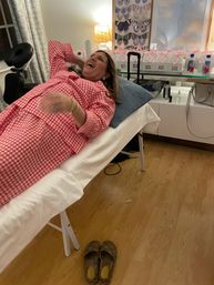 Laughing woman in pink gingham pajamas reclining on a white treatment table in a cozy home spa room with butterfly wall art, gift boxes, and slippers on a wood floor.