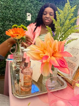 Vanity-style perfume display with a large peach-pink dahlia and orange gerbera, glass spray bottles on a mirrored tray, green foliage, and a blurred person in a pink robe against a leafy backdrop.