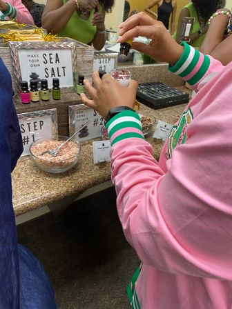 Hands pouring essential oil into a jar of DIY sea-salt scrub at a spa counter, with bowls of pink Himalayan salt and labeled oil bottles nearby
