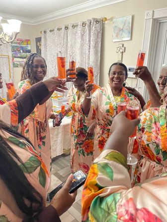 Women in matching floral satin robes smiling and raising tall red cocktails in a joyful at-home toast around a buffet table