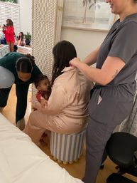 Woman in pink satin pajamas sits on a striped ottoman holding a toddler while a person in gray scrubs gives a shoulder massage at a cozy indoor pajama party with other guests in the background.